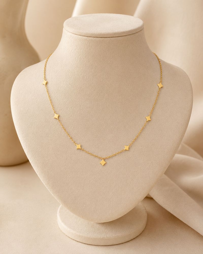 Collar BRÍLEA de NYXIDIE: cadena fina con estrellas en acero 304L acabado en oro de 18K, sobre busto de terciopelo marfil.